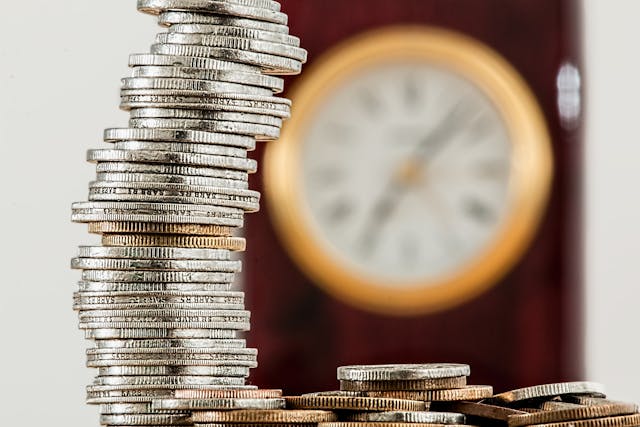 pexels-pixabay-128867 A stack of coins in front of a blurred out clock in the background